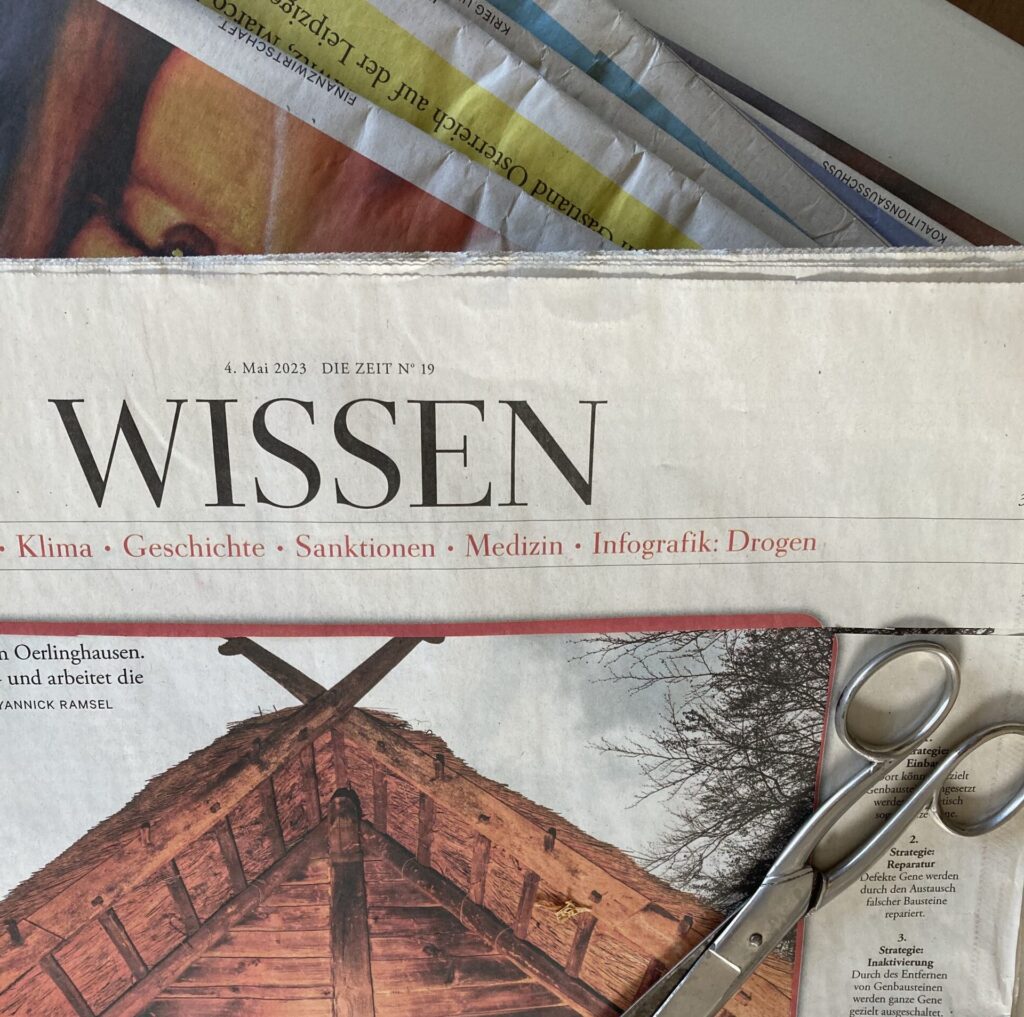 Eine Schere liegt auf einer umgedrehten Zeitung mit sichtbarem Text und einem Teilbild einer Holzkonstruktion. Darunter sind weitere Zeitungen teilweise sichtbar.